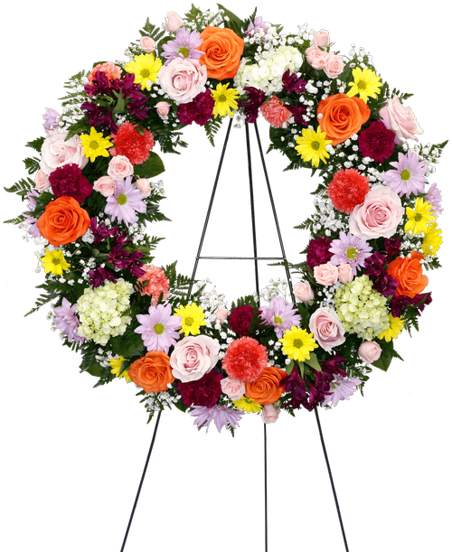 A wreath, suitable to be sent to a funeral or memorial service, is designed with a colorful mix of flowers including roses, carnations, alstroemeria, daisy poms, mini green hydrangea, and baby's breath. 26 inchD - Displayed on a 60 inch Easel