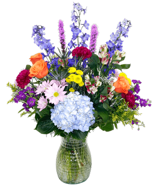 An 10 inch pebbled glass vase holds an all-around arrangement with three roses, delphinium, hydrangea, eryngium, charmelia alstroemeria, stock, carnations, daisy poms, button poms, liatris, solidago, and caspia. 31 inchH x 21 inchW