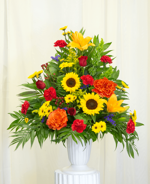 A one-sided arrangement from our Tuscan Funeral Collection, suitable to be sent to a funeral or memorial service, with roses, sunflowers, lilies, bells of Ireland, viking poms, carnations, hypericum, statice and solidago designed in a 11 1/4 inch urn. 33 inchH x 26 inchW