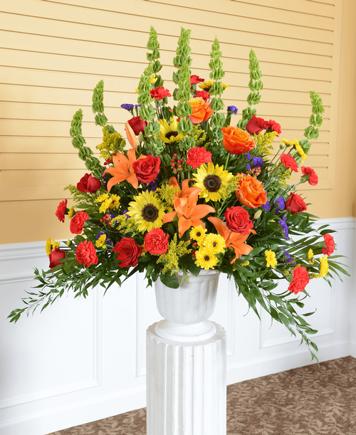 A one-sided arrangement from our Tuscan Funeral Collection, suitable to be sent to a funeral or memorial service, with roses, sunflowers, Bells of Ireland, lilies, viking poms, carnations, hypericum, statice and solidago designed in a 9.5 inch urn. 40 inchH x 36 inchW
