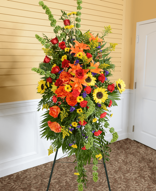 A memorial spray from our Tuscan Funeral Collection, with roses, sunflowers, lilies, carnations, Bells of Ireland, viking poms, hypericum, statice, and solidago. Displayed on a 60 inch Easel. 69 inchL x 33 inchW x 11 inchD