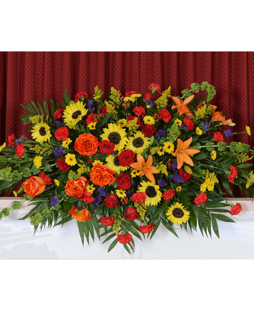 A casket spray from our Tuscan Funeral Collection with roses, sunflowers, lilies, carnations, Bells of Ireland, viking poms, hypericum, statice, and solidago. 72 inchL x 37 inchW x 13 inchH
