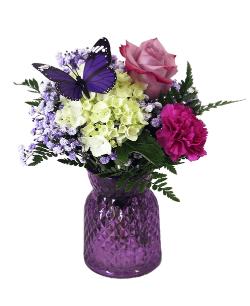 A 5.5 inchH lavender glass vase with a diamond pattern holds an all-around arrangement with a mini green hydrangea, a lavender carnation, a lavender rose, lavender daisy poms, purple-dyed baby's breath, and includes a butterfly stick-in. Overall 12 inchH x 7.5 inchW