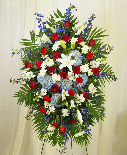 A standing spray from our Patriotic Funeral Collection, suitable to be sent to a funeral or memorial service, with roses, hydrangea, lilies, delphinium, stock, carnations, cushion poms, eryngium, and assorted greens. 54 inchL x 36 inchW - Displayed on a 54 inch Easel