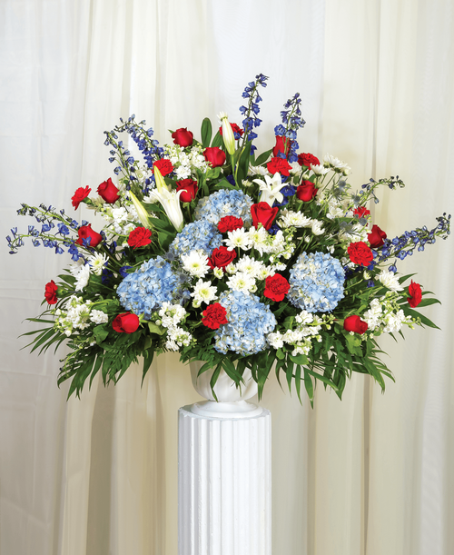 A one-sided arrangement from our Patriotic Funeral Collection, suitable to be sent to a funeral or memorial service, with roses, hydrangea, lilies, delphinium, stock, carnations, cushion poms, eryngium, and assorted greens designed in a 9.5 inch pedestal urn. 36 inchH x 38 inchW
