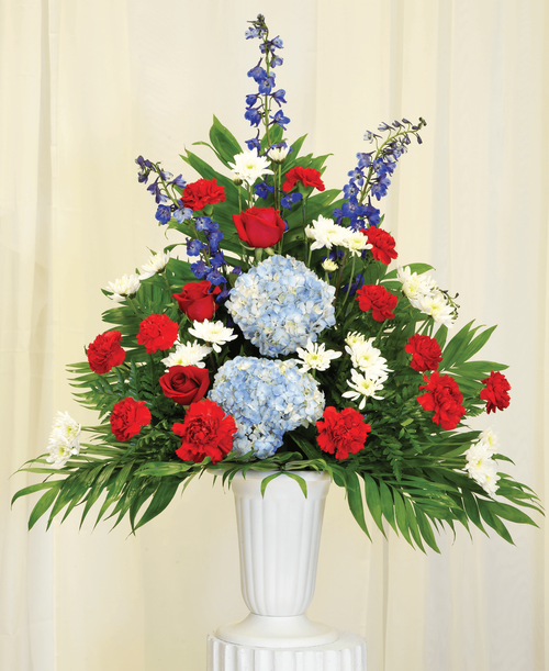 A one-sided arrangement from our Patriotic Funeral Collection, suitable to be sent to a funeral or memorial service, with roses, hydrangea, delphinium, carnations, cushion poms, and assorted greens designed in a 11-1/4 urn. 38 inchH x 28 inchW