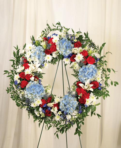 A 22 inch wreath from our Patriotic Funeral Collection, suitable to be sent to a funeral or memorial service, is designed with roses, hydrangea, delphinium, stock, lilies, carnations, cushion poms, eryngium, and assorted greens. Overall 33 inchD - Displayed on a 54 inch Easel