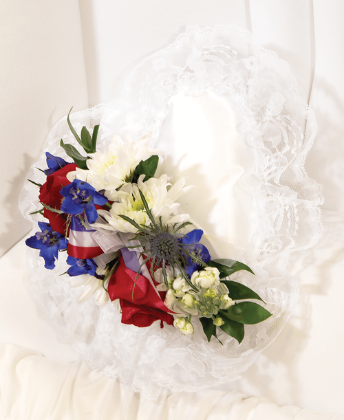 A 9 inch white pillow from our Patriotic Funeral Collection, suitable to be displayed at a funeral service inside the casket, is decorated with two roses, delphinium, stock, cushion poms, and eryngium. Cluster 10 inchL x 4 inchW