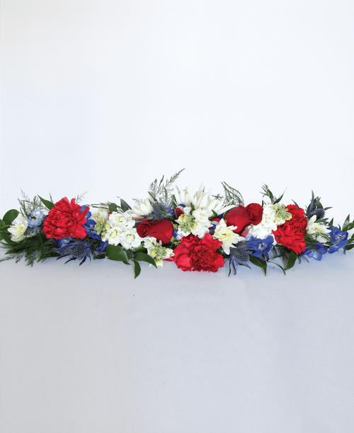 A ledgepiece from our Patriotic Funeral Collection to be placed inside the casket and suitable to be displayed at a funeral or memorial service, is designed with roses, carnations, stock, delphinium, carnations, cushion poms, a football mum, eryngium, and greens. 24 inchL x 7 inchW