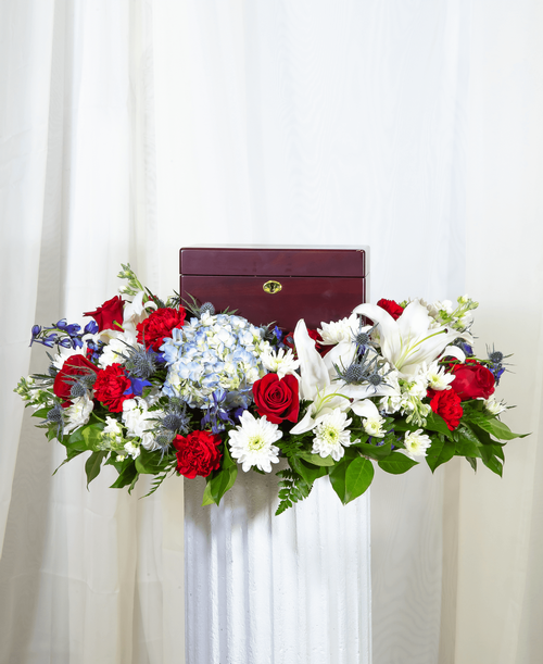 A memorial arrangement from our Patriotic Funeral Collection with roses, lilies, hydrangea, delphinium, stock, carnations, cushion poms, and assorted greens. Suitable for a funeral or memorial service with the option to display an urn, photograph or a special memento. 7 inchH x 28 inchD