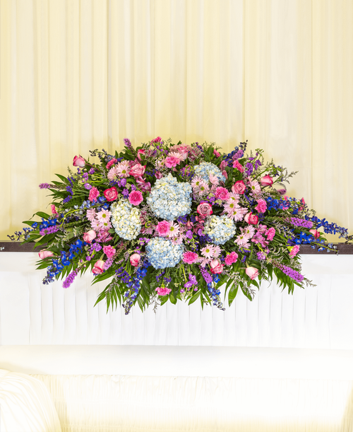 A full casket spray from our Tranquil Funeral Collection with roses, hydrangea, delphinium, stock, liatris, alstroemeria, carnations, daisy poms, statice, caspia, and assorted greens. 66 inchL x 30 inchW