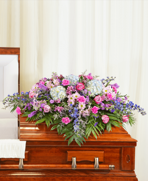 A half casket spray from our Tranquil Funeral Collection with roses, hydrangea, delphinium, stock, liatris, alstroemeria, carnations, daisy poms, statice, caspia, and assorted greens. 56 inchL x 50 inchW