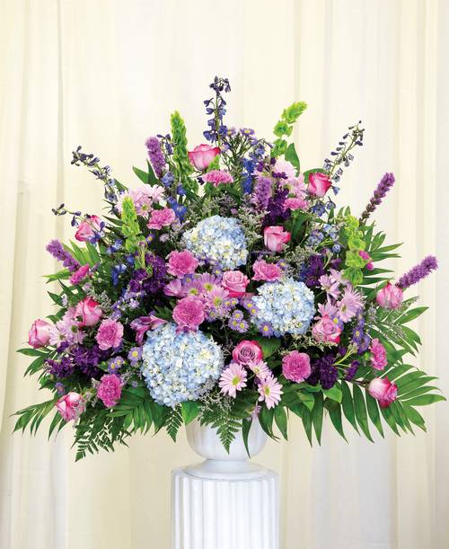 A one-sided arrangement from our Tranquil Funeral Collection, suitable to be sent to a funeral or memorial service, with roses, hydrangea, delphinium, stock, liatris, bells of Ireland, alstroemeria, daisy poms, statice, caspia, and assorted greens designed in a 9.5 inch pedestal urn. 38 inchH x 36 inchW