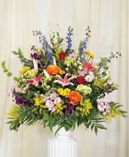 A one-sided arrangement from our English Garden Funeral Collection, suitable to be sent to a funeral or memorial service, with roses, hydrangea, delphinium, stock, lilies, alstroemeria, bells of Ireland, daisy poms, viking poms, solidago, caspia, and assorted greens designed in a 9.5 inch pedestal urn. 38 inchH x 36 inchW; Overall 40 inchH