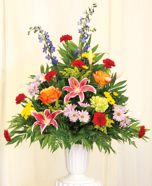 A one-sided arrangement from our English Garden Funeral Collection, suitable to be sent to a funeral or memorial service, with lilies, mini hydrangea, delphinium, carnations, daisy poms, viking poms, solidago, and curly willow accents designed in a 11 1/4 inch urn. Overall 38 inchH x 28 inchW