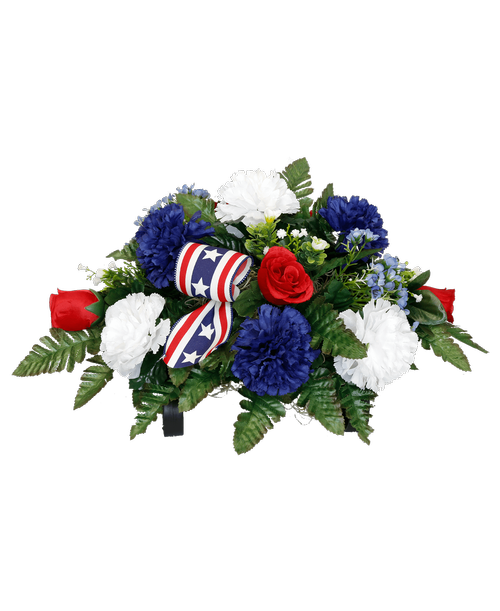 A cemetery gravestone saddle holds a one-sided silk arrangement with red, white, and blue flowers including carnations, roses, waxflower, and two bows. 18 inchL x 14 inchW x 9.5 inchH (flowers)